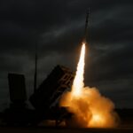 A Patriot missile defense system launches a surface-to-air missile into a stormy sky, with bright flames illuminating the clouds and silhouetted radar arrays in the background.