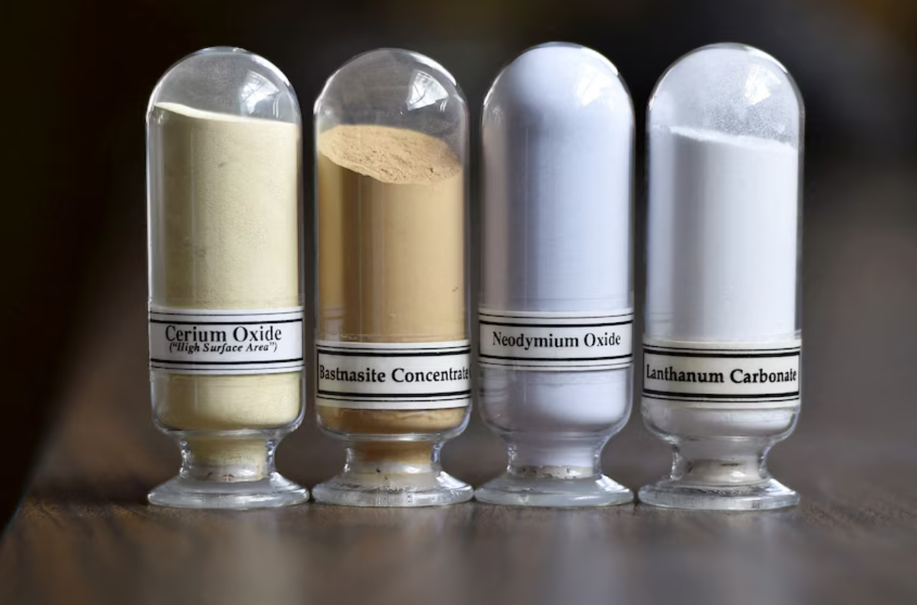 Samples of refined rare earth minerals including cerium oxide, bastnasite concentrate, neodymium oxide, and lanthanum carbonate displayed in glass containers, symbolizing their role in AI, medical technology, renewable energy, and defense industries